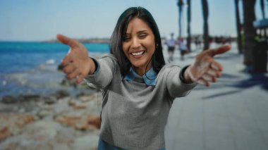 Okyanus manzaralı deniz kıyısında gülümseyen ve kollarını uzatan genç bir kadın açık havada neşeli bir manzara yakalıyor..