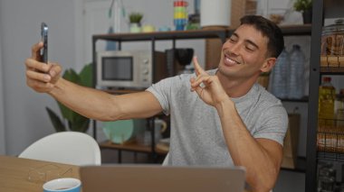 Modern ofis ortamında gülümseyen, pozitiflik ve rahatlamış atmosfer aktaran genç İspanyol bir adam, bir video çağrısı sırasında barış jesti yapıyor..