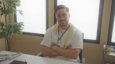 Doktor kıyafeti ve steteskop giyen sakallı genç adam kollarını kavuşturup klinik ortamında profesyonel bir sağlık hizmeti sunuyor..