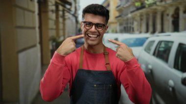 Önlüklü genç adam, şehir sokaklarında günlük kıyafetleriyle dikilirken dişlerini göstererek şehir ortamında neşeli ve dostane bir tavır sergiliyor..