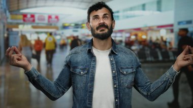 Sakallı İspanyol bir adam havaalanı terminalinde meditasyon yapıyor. Etrafı kalabalık bir atmosferde yolcularla çevrili. Modern terminalde seyahat kaosunun ortasında huzurlu bir an yaratıyor..