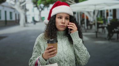 Dışarıda Noel Baba şapkası takan bir kadın, elinde kahve poşetleri ve alışveriş poşetleri tutarken tatil sezonunda şehrin bir caddesinde akıllı bir telefonla konuşurken, şenlikli bir şehir ortamının altını çiziyor..