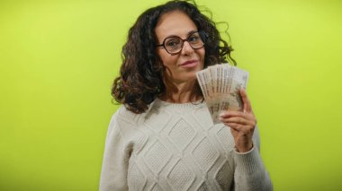 Orta yaşlı bir kadın, sade sarı bir duvarın önünde Rus rublesi tutuyor. Parlak, izole bir ortamda kendine güvenen bir ifadeyle para ve servet sergiliyor..