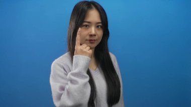 Hırkalı genç bir kadın mavi arka plan duvarının üzerinde parmak göze işaret ediyor..