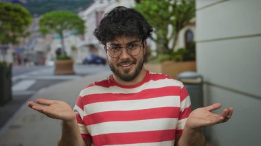 Sakallı genç adam canlı bir sokak ortamında gülümsüyor, gözlük takıyor ve kırmızı çizgili bir gömlek giyiyor, mutluluk ve rahatlama taşıyor..