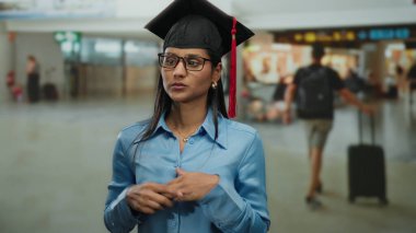 Mezuniyet şapkalı ve gözlüklü genç bir kadın havaalanı terminalinde duruyor. Arka planda bulanık gezginler geçişi ve yeni başlangıçları simgeliyor..