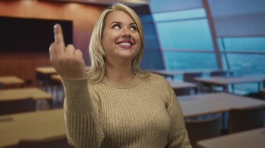 Gülümseyen genç sarışın kadın, ahşap masaları ve geniş pencere manzarasıyla sınıfta kamerayı işaret ederek, coşkuyu öğreniyor..