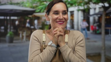 Önlüklü İspanyol kadın canlı bir sokakta kafeler ve ağaçlarla neşeyle tepki veriyor. Canlı bir şehir ortamında şaşkınlık ve keyif ifade ediyor..