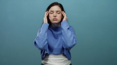 Esmer saçlı bir kadın, mavi bir arka planda stresli hissediyor. Mavi bir kazak giyip baş ağrısıyla boğuşuyor. Duyguları ve gerilimi canlı bir şekilde yakalıyor..