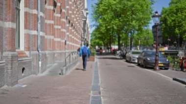 Bulanık bir adamın güneşli bir günde Avrupa şehir kaldırımında yürüdüğü şehir sokak sahnesi klasik mimari ve arabalarla park edilmiş, canlı bir açık hava havası yakalamış..