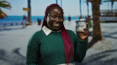 Deniz kenarındaki gezinti güvertesinde elinde dolar banknotuyla cam kavanoz tutan, yeşil kazak giyen, güneşli sahil arka planına karşı duran, kırmızı örgülü ve açık havada okyanusa bakan bir kadın..