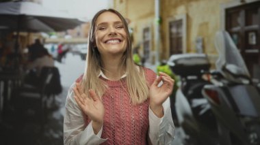 Bir kadın açık hava restoranının terasında el çırpıyor, gündüz vakti sokak manzarasının ortasında gülümsüyor..