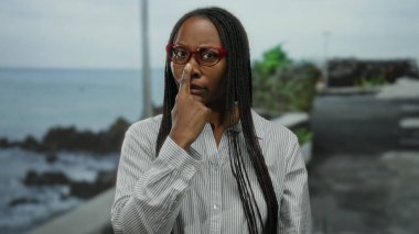 Gözlüklü kadın, deniz kenarında bir gezinti güvertesinde neşeli bir şekilde gülümsüyor. Okyanusun sakin arka planına karşı enerjik bir enerji ve ifade sergiliyor..