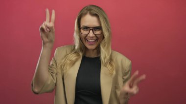 Kırmızı arka planda barış işaretleriyle poz veren sarı saçlı ve gözlüklü bir kadın..