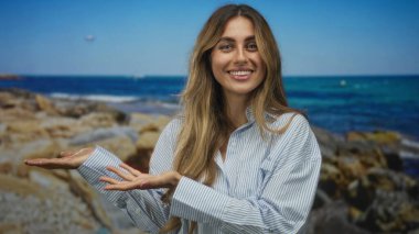 Gülümseyen genç sarışın kadın kayalık deniz kıyısında avuçlarını açık tutuyor; davetkar keşif huzur neşe özgürlüğü.