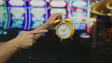 Kumarhanede sarı saat tutan beyaz bir adam. Arka planda kumar makineleri var. Zamana ve kumar atmosferine önem veriyor..