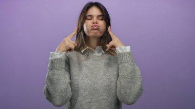 Latin kökenli genç bir kadın mor bir arka plana karşı neşeli bir yüz ifadesi takınıyor, rahat bir süveter giyiyor ve pofuduk yanaklarıyla ve neşeli hareketleriyle duygularını ifade ediyor.