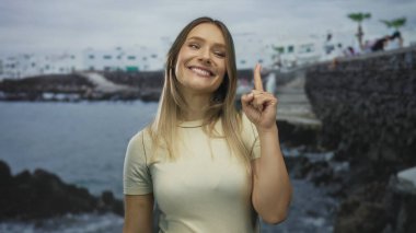 Sahilde gülümseyen bir kadın sahilde arka planda beyaz binaları işaret ederek bulutlu bir gökyüzünün altında açık denizde neşeli bir ifade sergiliyor..