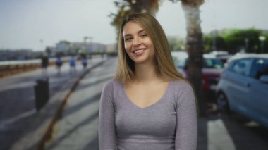 Güneşli bir sahil gezisinde, park etmiş arabalarla ve bulanık denizlerle gülümseyen bir kadın açık havada zarafetini sergiliyor..