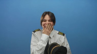 Heyecanlı İspanyol kadın pilot mavi arka planda şapkasını tutuyor şaşırmış ifadesiyle ve izole edilmiş duvar düzeniyle