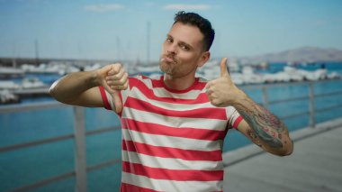 Sahil limanındaki çizgili gömlekli genç adam, açık havada karışık duygular olduğunu düşündüren tekne ve su arkaplanlarına karşı dövmeleri olan baş parmaklarını hem yukarı hem aşağı gösteriyor.