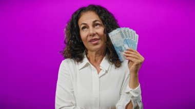 Orta yaşlı bir kadın canlı mor bir duvara karşı Şili pezosu tutuyor. Düşünceli bir ifadeyle para ve bankacılık temalarını vurguluyor..