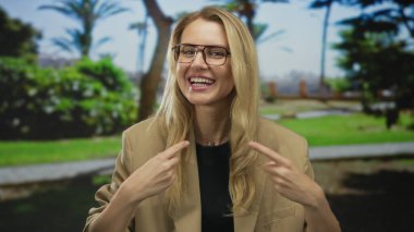 Güneşli bir parkta gülümseyen ve kendini gösteren bir kadın. Etrafı yemyeşil, siyah tişörtlü ve gündüz gözlüklü bej ceketli..