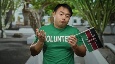 Yeşil gönüllü gömlekli genç Asyalı bir adam kenya bayrağını arka planda ağaçların oluşturduğu bir şehir ortamında tutuyor..