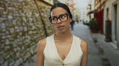 Dar bir sokak arkaplanında tuhaf bir ifadeyle, gözlüklü kadın, farklı bir Avrupa tarzında kentsel hayat sergiliyor, modern sokak modasını yansıtıyor..