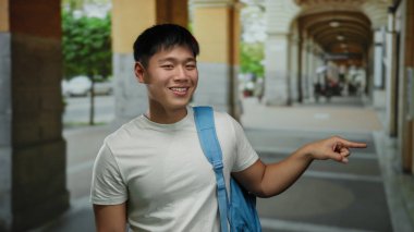 Sırtında mavi bir sırt çantasıyla şehir yaşamını gözler önüne seren, gülümseyen ve işaret eden genç bir adam..