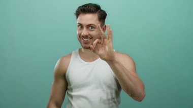 Beyaz askılı sakallı genç adam yeşil bir arka plana gülümseyen, dışa dönük, dışa dönük yüz ifadelerini sergileyen iyi bir işaret veriyor..