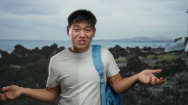 Sırt çantalı genç adam, kayalık sahil kenarında el kol hareketi yapıyor. Sahilde rahat bir öğrenci havası var. Bulutlu bir günde açık havada, deniz kenarında bir anı yakalıyor..