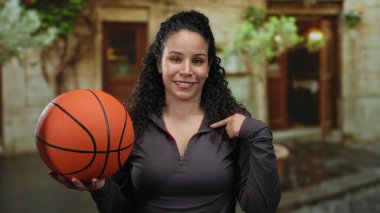 Elinde basketbolla kenar mahalle ortamını işaret eden bir kadın, arka planı bulanık ve rahat bir şekilde gülümseyen genç bir İspanyol..