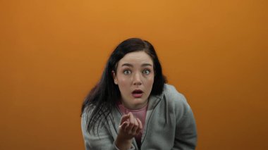Ağzı açık şaşırmış bir kadın gri kapüşonlu giyer parlak turuncu arka planda durur şok ve duygu taşır..