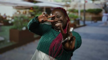 Kırmızı örgülü gülümseyen kadın bir restoranın dışındaki terasta zafer jesti yapıyor..