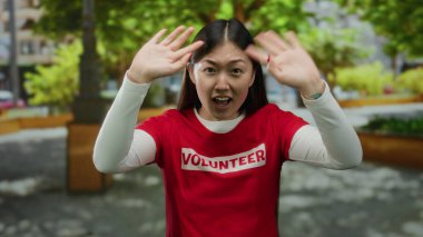 Gönüllü gömleği giyen, kolları parkta yükselen, arka planda ağaçlar ve binalar olan genç bir kadın, coşku ve açık havada toplum ruhunu gösteriyor..