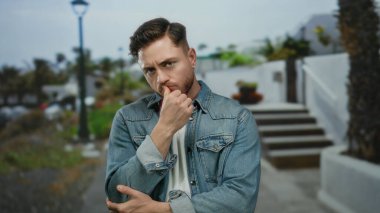 Sakallı genç bir adam, şehir dışında bir sokakta kot ceket giyerek derin derin düşünüyor..