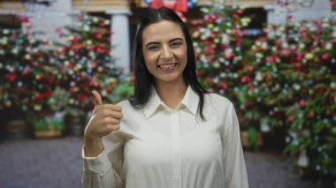 Açık hava ortamında bulanık arkaplanı olan neşeli caddede baş parmaklarıyla gülümseyen kadın, tatil sezonunda mutluluk ve pozitifliği işaret ediyor..