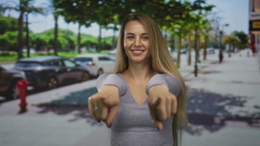 İki eliyle işaret eden ve arka planda arabalarla dolu güneşli bir şehir caddesinde gülümseyen bir kadın ve yürüyen insanlar, canlı bir şehir atmosferi yaratıyor..