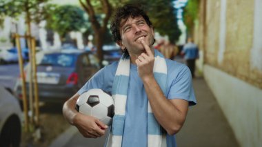 Hispanik bir adam sokakta, elinde düşünceli bir ifadeyle futbol topu tutuyor, şehir sporu hevesini somutlaştırıyor..