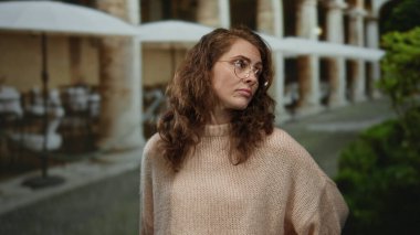 Şehir restoranındaki terasta duran bir kadın ilgisizliğini dile getiriyor, süveter giymiş, masa ve şemsiyelerden oluşan bir arka planı var, şehir havası yayıyor..