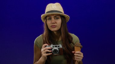 Kameralı ve dondurmalı genç bir kadın, izole edilmiş mavi bir arka plan duvarına karşı duruyor. Canlı bir ortamda, bir yaz şapkası takmış, bir macera ve tatlılık anı yakalıyor..
