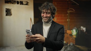 İnşaat alanında elinde akıllı telefon tutarken gülümseyen yakışıklı İspanyol adam. Arka planda bitmemiş duvarlar, şık bir siyah takım elbise ve bej balıkçı yaka giyiyor.