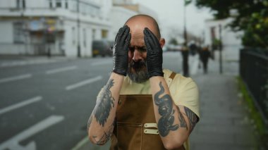 Sakallı, eldiven ve önlük takan, şehir caddesinde dikilen, elleriyle el kol hareketi yapan bir adam..
