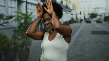 Gülümseyen bir kadın, şehir sokaklarında elleri ile neşeli bir jest yaparak, açık şehir ortamında neşe ve pozitifliği canlı bir enerjiyle vurguluyor.
