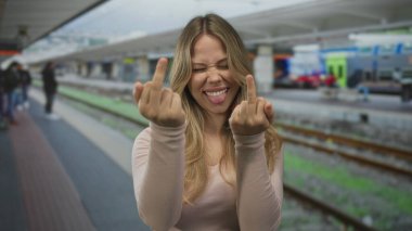 Genç beyaz kadın, kalabalık tren istasyonunda orta parmaklarını sallıyor ve dilini dışarı çıkarıyor..