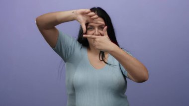 Bir kadın, açık mavi bluz giyerek, elleri izole edilmiş mor arkaplan üzerinde bir çerçeve jesti yapıyor, oyuncu bir duruşta yaratıcılık ve hayal gücünü ifade ediyor..