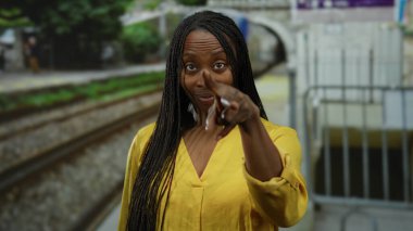 Sarı elbiseli kadın açık hava tren istasyonunda duruyor. Mesajı parmağıyla, tren ve raylarıyla gösteriyor. Dinamik bir şehir ulaşımı sahnesi yaratıyor..
