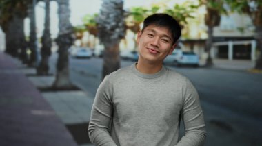 Palmiye ağaçları ve bulanık şehir arkaplanı ile dolu bir sokakta gelişigüzel gülümseyen genç adam, doğal ve insan yapımı elementlerin şaşırtıcı bir karışımı ile canlı bir şehir atmosferi yaratıyor..