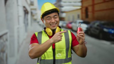 Güler yüzlü Asyalı adam emniyet yeleği giyiyor ve kentsel caddedeki elektronik cihaza nokta koyuyor. Arka planda arabaları var. Şehir temalı açık hava portresi çizimleri yapıyor.